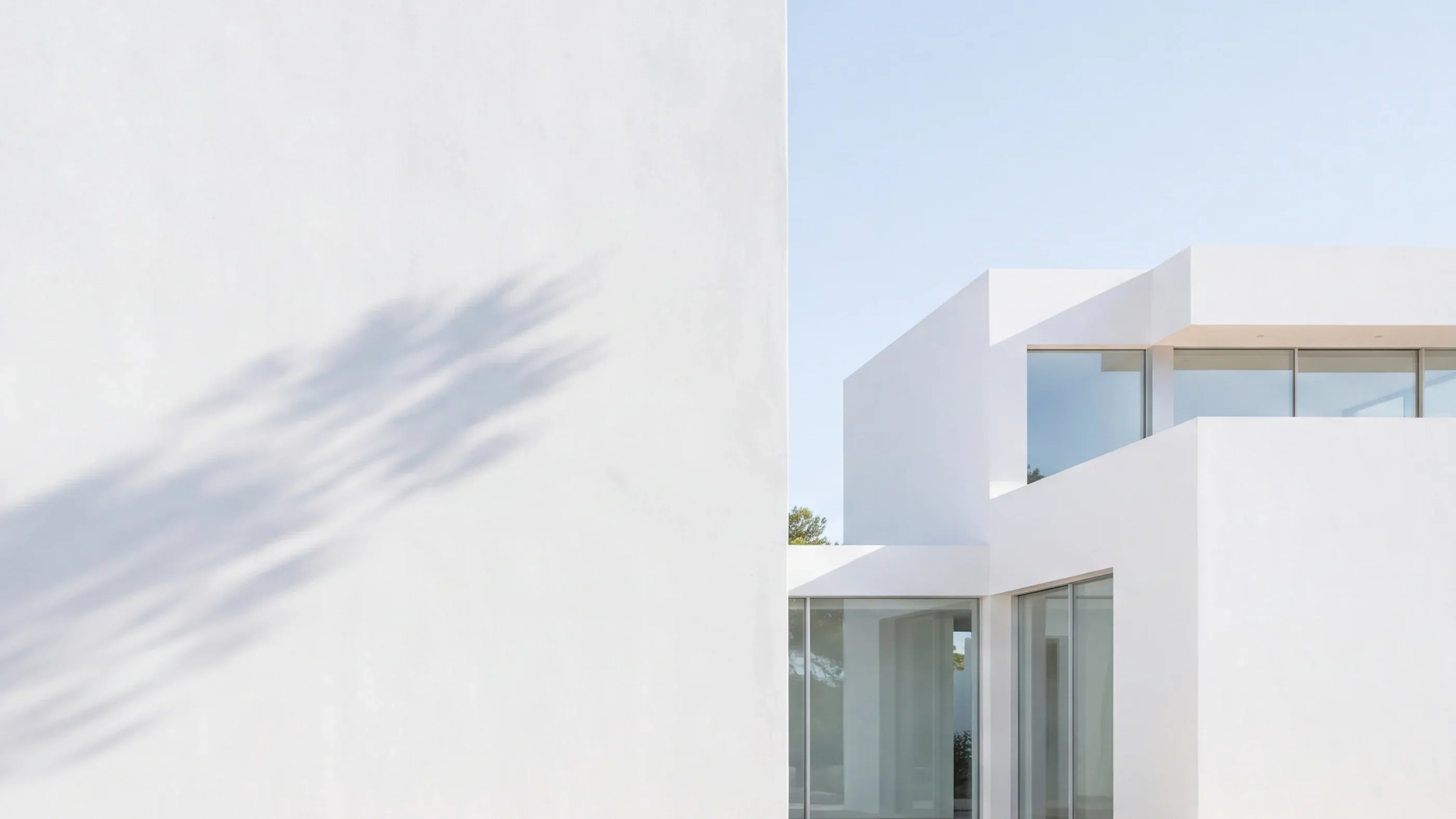 White cubic volumes of a modern Ibizan villa in bright daylight, next to a bare rendered wall crossed by the shadow of a pine